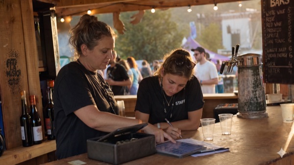 Zwei Helferinnen bei der Schichtübergabe hinter der Festival-Bar, eine zeigt etwas auf einem Clipboard